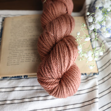 A Bulky sparkle skein of yarn colored  a moody salmon pink tonal-solid, sitting on top of a book. there is a blue and white vase with white flowers  sticking out of it. All are sitting on top of a black and white striped table cloth.