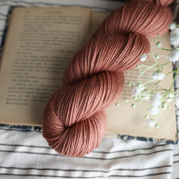 A DK skein of yarn colored  a moody salmon pink tonal-solid, sitting on top of a book. There are white flowers hovering over the skein. All are sitting on top of a black and white striped table cloth.