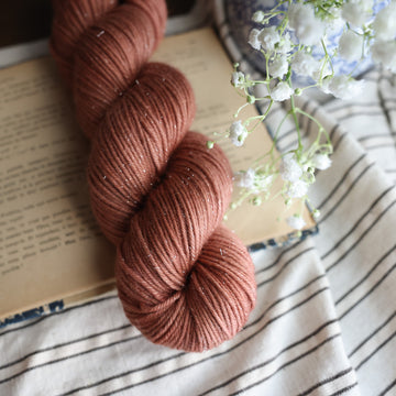 A DK sparkle skein of yarn colored  a moody salmon pink tonal-solid, sitting on top of a book. there is a blue and white vase with white flowers  sticking out of it. All are sitting on top of a black and white striped table cloth.