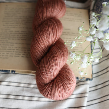 A fingering sparkle skein of yarn colored  a moody salmon pink tonal-solid, sitting on top of a book. there is a blue and white vase with white flowers  sticking out of it. All are sitting on top of a black and white striped table cloth.