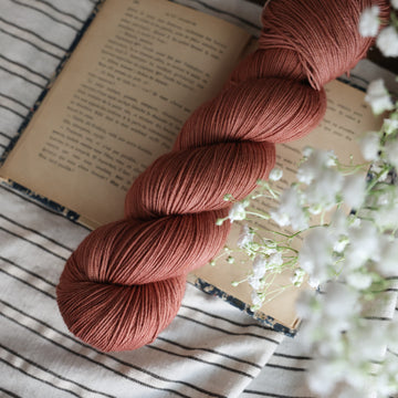 A fingering skein of yarn colored  a moody salmon pink tonal-solid, sitting on top of a book. There are white flowers hovering over the skein. All are sitting on top of a black and white striped table cloth.