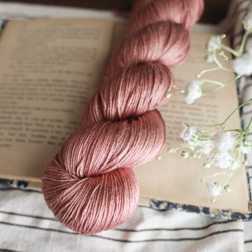 A silk skein of yarn colored  a moody salmon pink tonal-solid, sitting on top of a book. There are white flowers hovering over the skein. All are sitting on top of a black and white striped table cloth.