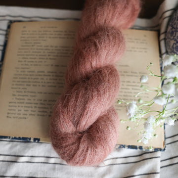 A Suri Alpaca skein of yarn colored  a moody salmon pink tonal-solid, sitting on top of a book. there is a blue and white vase with white flowers  sticking out of it. All are sitting on top of a black and white striped table cloth.