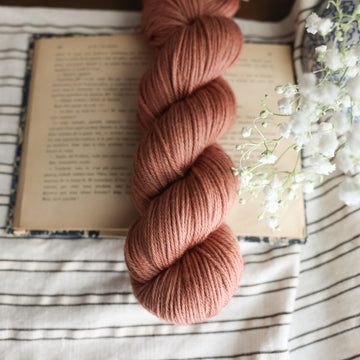 A Worsted skein of yarn colored  a moody salmon pink tonal-solid, sitting on top of a book. there is a blue and white vase with white flowers  sticking out of it. All are sitting on top of a black and white striped table cloth.