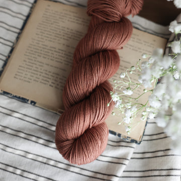 A sport skein of yarn colored  a moody salmon pink tonal-solid, sitting on top of a book. There are white flowers hovering over the skein. All are sitting on top of a black and white striped table cloth.