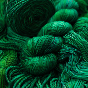 An emerald green Sport yarn that is on top of the same colored unwound yarn in different weights and a caked version of the yarn on the side.