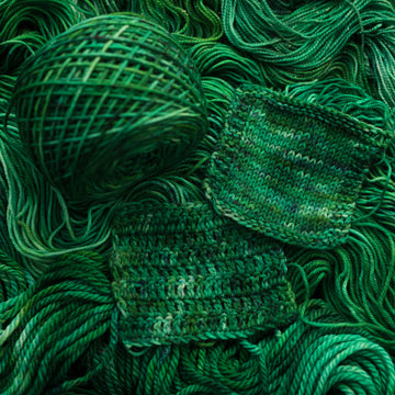Emerald green colored knit and crochet swatches that is on top of the same colored unwound yarn in different weights.