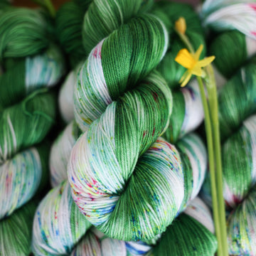 Wound up Fingering yarn that is colored with a darkish green with some lighter shades mixed in, and a white streak coming through the green on both sides that has different colored speckles with light greens pinks and blues. There are yellow flowers on top of the yarn.