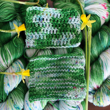 Wound up yarn with the same color yarn in crochet and knit swatches on top that is colored with a darkish green with some lighter shades mixed in, and a white that has different colored speckles with light greens pinks and blues. There are yellow flowers on top of the yarn.
