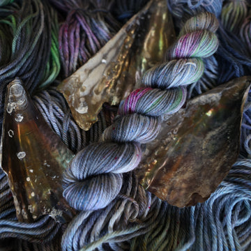 Fingering mini yarn colored with different shades of grey and streaks of green, and dark pink, also there are also blue and purple speckles. These skeins are on top of unwound skeins of the same color way with some shells on top.