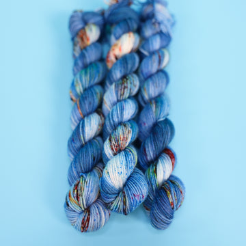 Fingering mini yarn colored with different shades of blue and white with multicolored speckles on the portion of the yarn that Is white. all on top of a light blue background.