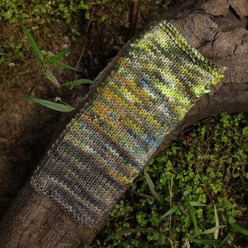 A large knitted swatch with three different color ways faded together, on top of a tree limb that is hovering over a pond, with different mossy plants around the pond, and around the yarn.