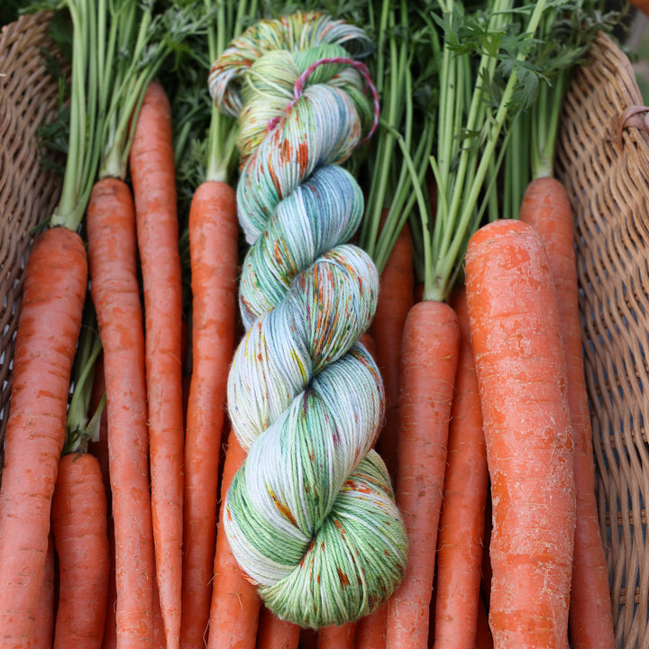 Silk yarn colored different shades of light and dark green, white, some blue, yellow, and speckles of orange red. This yarn is in a basket of garden carrots.