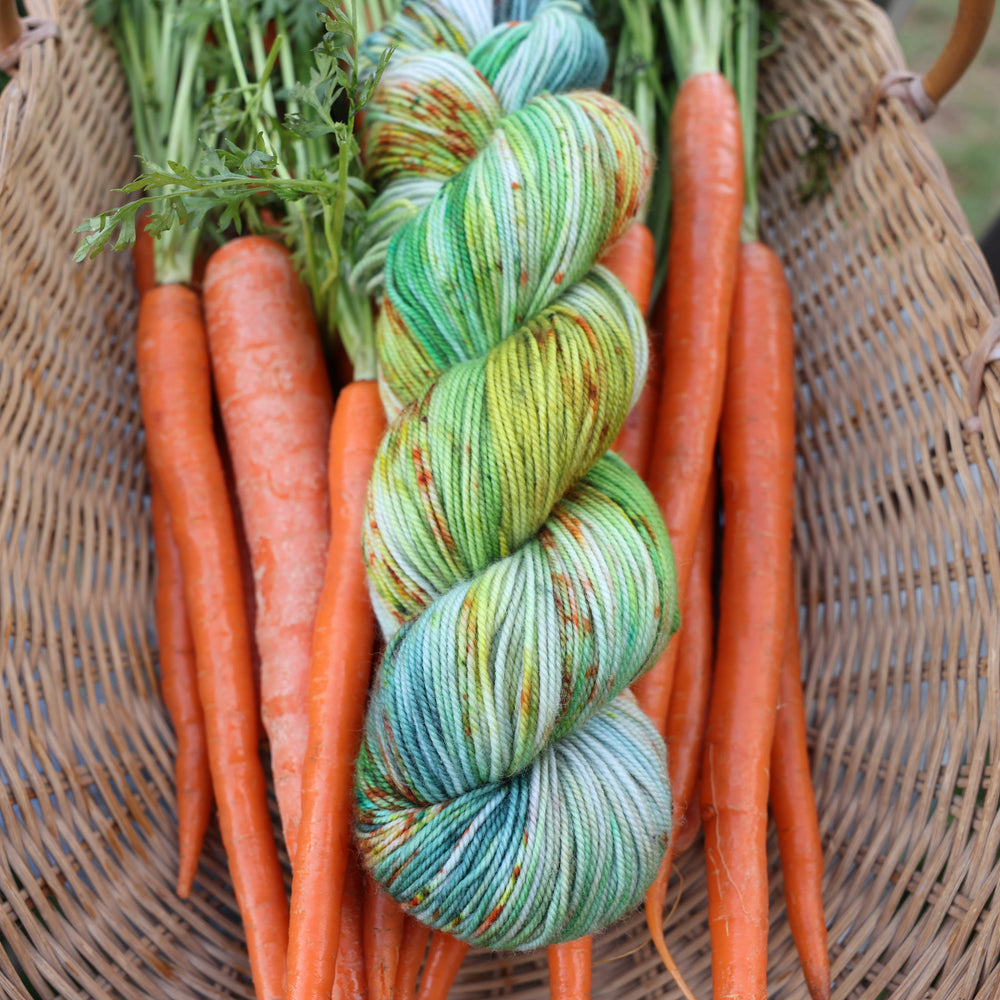 Sport yarn colored different shades of light and dark green, white, some blue, yellow, and speckles of orange red. This yarn is in a basket of garden carrots.