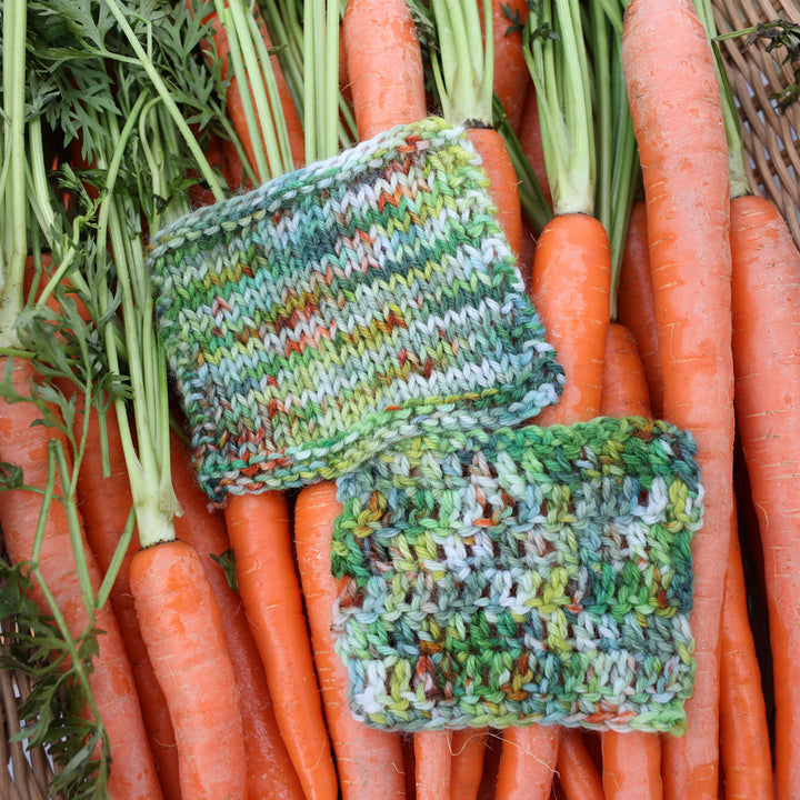 Crochet and knit swatches colored different shades of light and dark green, white, some blue, yellow, and speckles of orange red. These swatches are in a basket of garden carrots.
