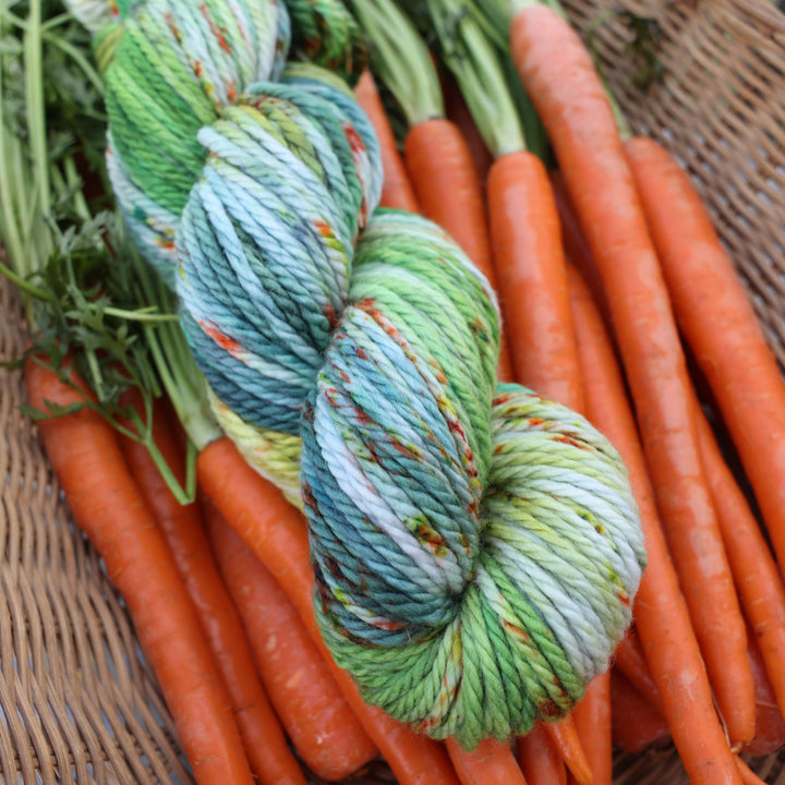Bulky yarn colored different shades of light and dark green, white, some blue, yellow, and speckles of orange red. This yarn is in a basket of garden carrots.