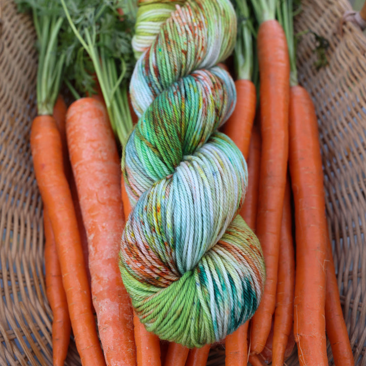 DK yarn colored different shades of light and dark green, white, some blue, yellow, and speckles of orange red. This yarn is in a basket of garden carrots.