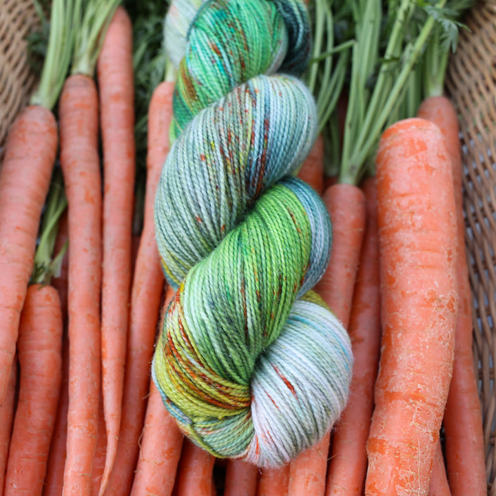 Sparkle fingering yarn colored different shades of light and dark green, white, some blue, yellow, and speckles of orange red. This yarn is in a basket of garden carrots.