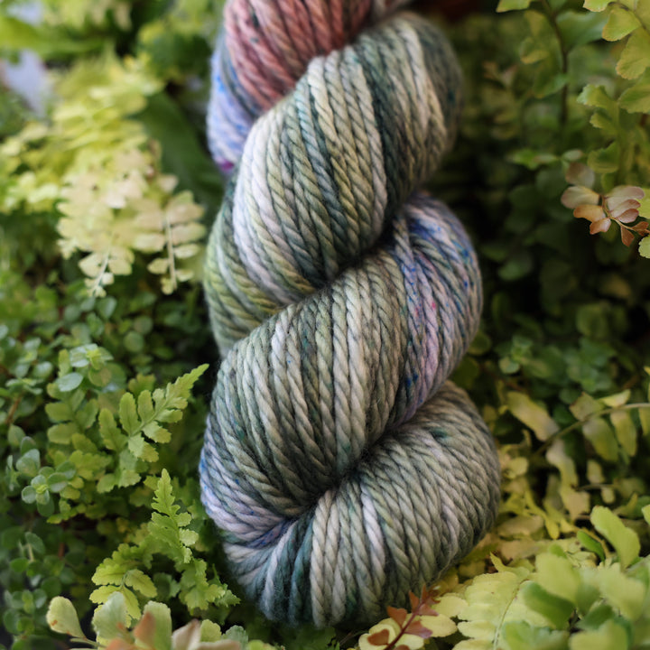 Bulky yarn colored different colors like forest green light pink, some blue green colors, and white and purple. This skein is on top of a bed of ferns and plants.