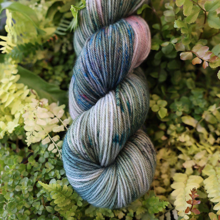 DK yarn colored different colors like forest green light pink, some blue green colors, and white and purple. This skein is on top of a bed of ferns and plants.