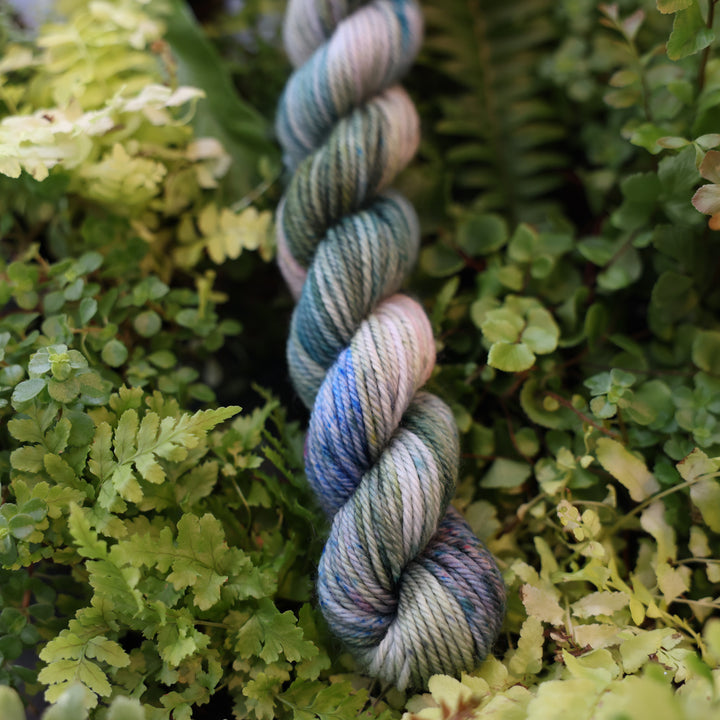 DK mini yarn colored different colors like forest green light pink, some blue green colors, and white and purple. This skein is on top of a bed of ferns and plants.