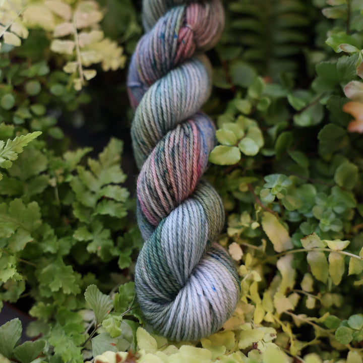 Fingering mini yarn colored different colors like forest green light pink, some blue green colors, and white and purple. This skein is on top of a bed of ferns and plants.