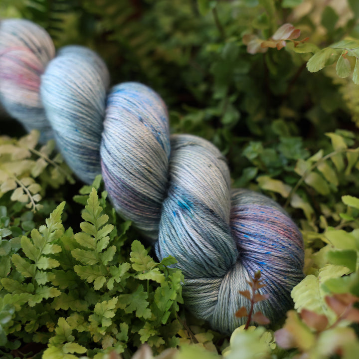 Silk yarn colored different colors like forest green light pink, some blue green colors, and white and purple. This skein is on top of a bed of ferns and plants.