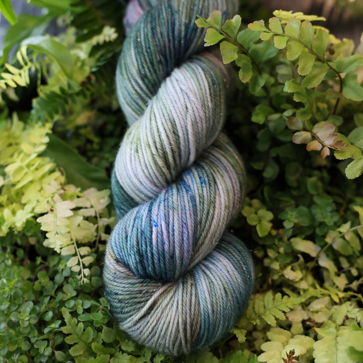 DK sparkle yarn colored different colors like forest green light pink, some blue green colors, and white and purple. This skein is on top of a bed of ferns and plants.