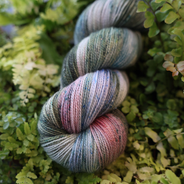 Sparkle fingering yarn colored different colors like forest green light pink, some blue green colors, and white and purple. This skein is on top of a bed of ferns and plants.