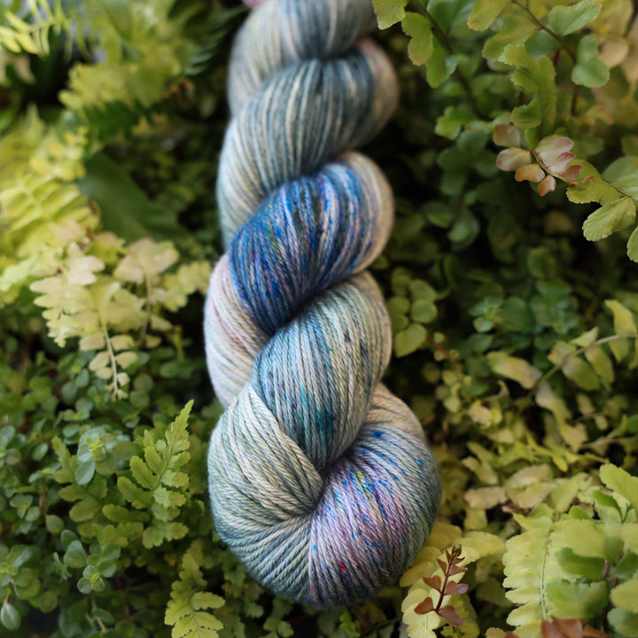 DK yarn colored different colors like forest green light pink, some blue green colors, and white and purple. This skein is on top of a bed of ferns and plants.