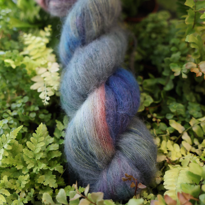 Suri alpaca yarn colored different colors like forest green light pink, some blue green colors, and white and purple. This skein is on top of a bed of ferns and plants.