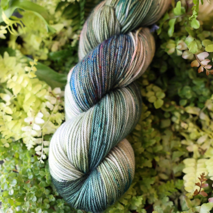 Sport yarn colored different colors like forest green light pink, some blue green colors, and white and purple. This skein is on top of a bed of ferns and plants.