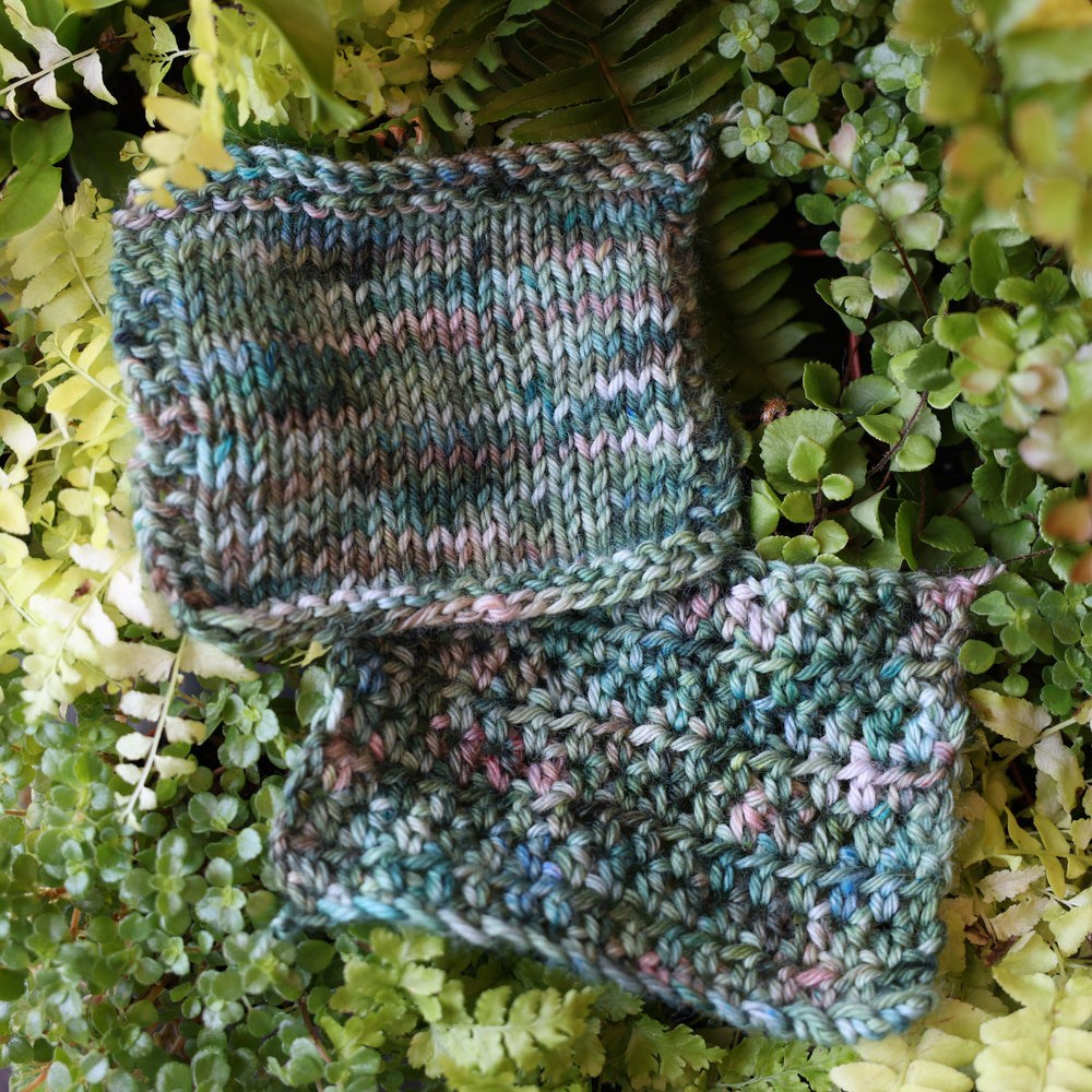 crochet and knit swatches colored different colors like forest green light pink, some blue green colors, and white and purple. These swatches are on top of a bed of ferns and plants.