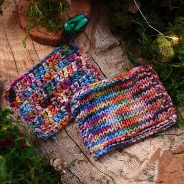 Knit and crochet swatches colored with a hot pink, light blue, dark blue/purple, with some green and orange/brown colors, on top of a wooden table, with moss in the corners, fairy lights, and a glass looking beetle on a wooden slab .