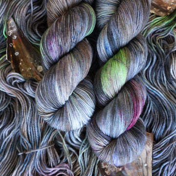 Fingering yarn colored with different shades of grey and streaks of green, and dark pink, also there are also blue and purple speckles. These skeins are on top of unwound skeins of the same color way with some shells on top.