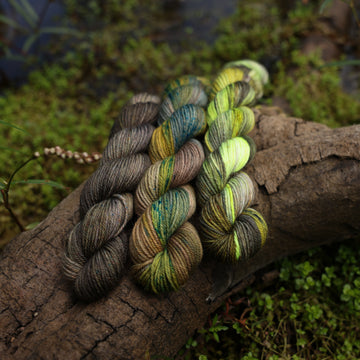 Different Fingering mini yarn skeins, one colored a lime green and grey, one colored a dark grey, and one colored green with blue speckles, on top of a tree limb that is hovering over a pond, with different mossy plants around the pond, and around the yarn.