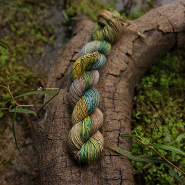A Fingering mini yarn skein colored with green, yellow and grey colors, with blue speckles, on top of a tree limb that is hovering over a pond, with different mossy plants around the pond, and around the yarn.