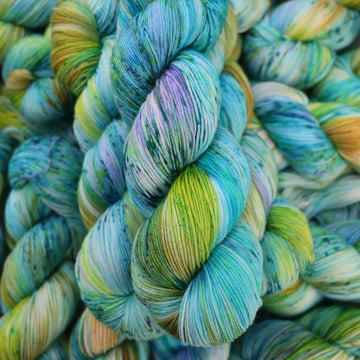 Fingering yarn with light shades of blue and speckles of purple, with some light greens and browns. There are more skeins like this of different weights underneath.