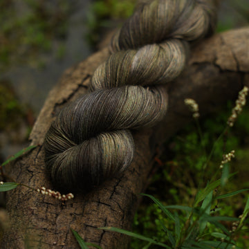 A dark grey Fingering yarn with hints of green colors, on top of a tree limb that is hovering over a pond, with different  mossy green plants around the pond, and around the yarn.