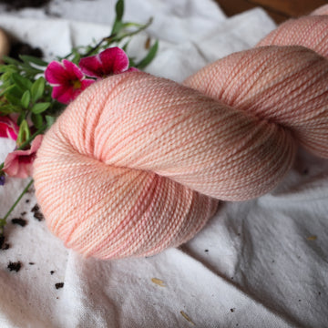 A light pink colored Fingering yarn skein on top of a white tablecloth with some pink flowers on a brown wooden table.