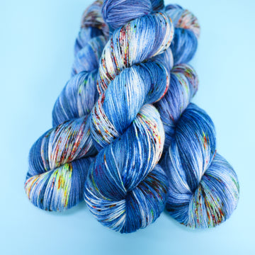 Fingering yarn colored with different shades of blue and white with multicolored speckles on the portion of the yarn that Is white. all on top of a light blue background.