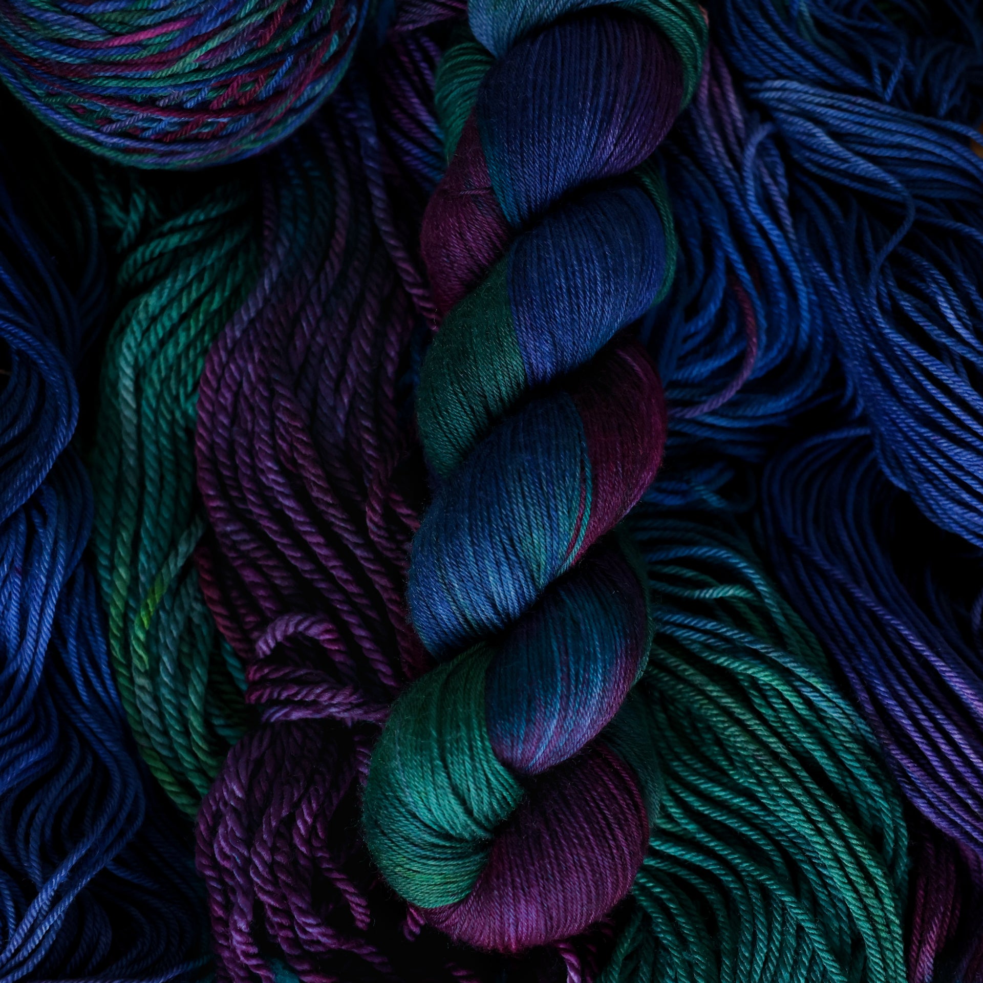 Fingering yarn colored with an emerald green, Dark blue, and dark purple, on top of un unwound version of that yarn in a different weight.
