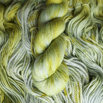 Fingering yarn colored with different shades of light green and white with some dark green speckles on top of the same unwound yarn.