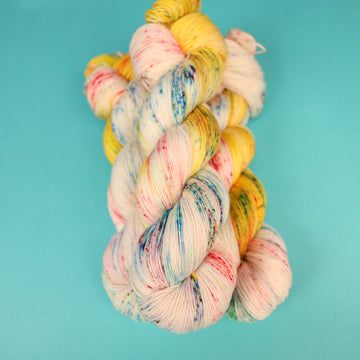 A light cream colored Fingering yarn with different colored speckles all over and banana colored yellow at the top of the skein on top of a light blue background.