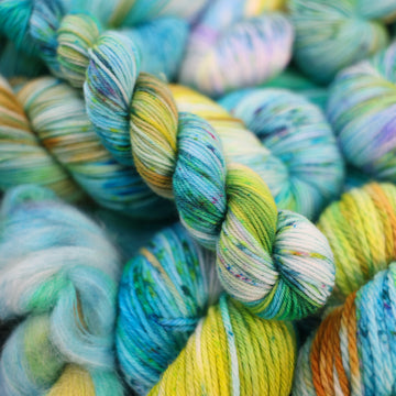 Fingering mini yarn with light shades of blue and speckles of purple, with some light greens and browns. There are more skeins like this of different weights underneath.