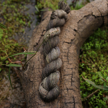 A dark grey fingering mini yarn with hints of green colors, on top of a tree limb that is hovering over a pond, with different plants around the pond, and around the yarn.