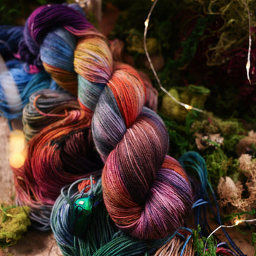 Fingering yarn colored with a hot pink, light blue, dark blue/purple, with some green and orange/brown colors, on top of the same yarn but unwound, with moss underneath and some fairy lights around the moss and a small glass looking beetle.