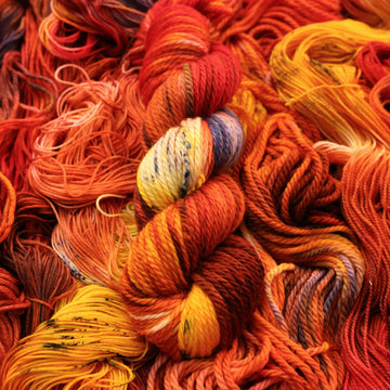 A bright red and orange colored Bulky yarn that has hints of purple and white and some yellow and brown colors. There are more weights of this yarn unwound underneath this skein.