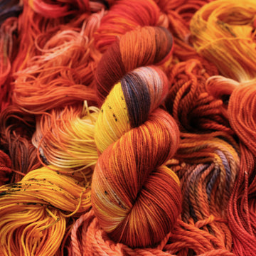 A bright red and orange colored Fingering 
yarn that has hints of purple and white and some yellow and brown colors. There are more weights of this yarn unwound underneath this skein.