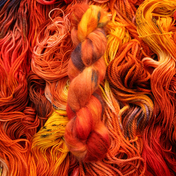 A bright red and orange colored Suri Alpaca yarn that has hints of purple and white and some yellow and brown colors. There are more weights of this yarn unwound underneath this skein.